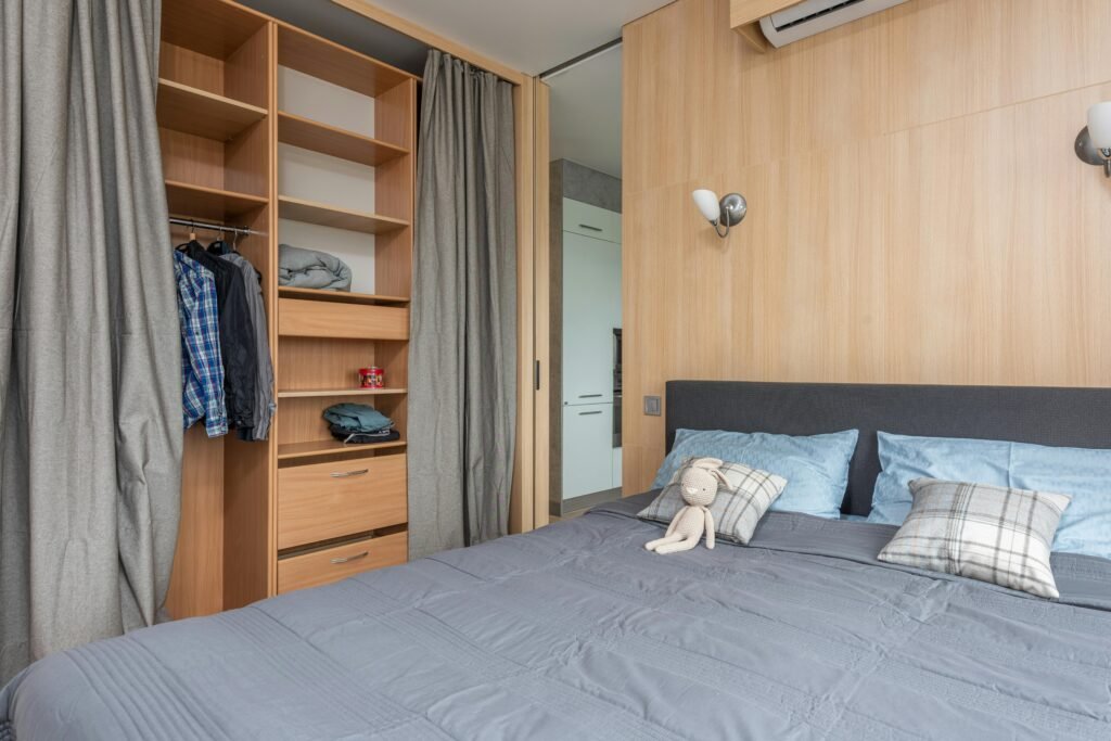 Modern room interior with bed with pillows and soft rabbit toy near wooden wardrobe with curtains and shelves near clothes on hangers and entrance to kitchen in bright flat
